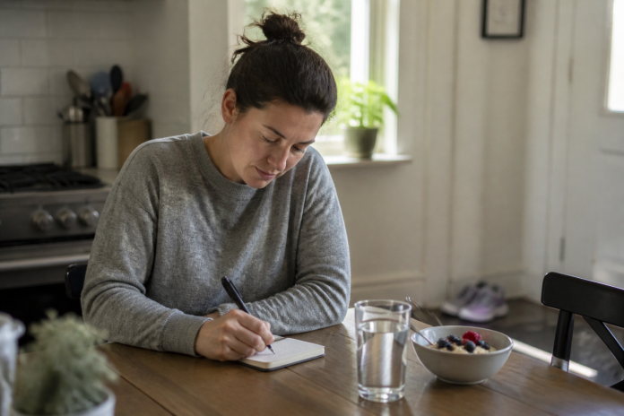 How to Live a Happy Life: Daily Basics That Actually Help Woman writing a short note in a notebook at a kitchen table in morning light, with water, breakfast and running shoes nearby
